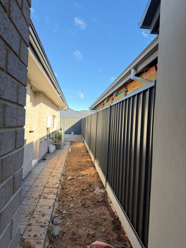 Narrow alleyway between two residential buildings with brick walls, metal fence, and bare ground under clear blue sky