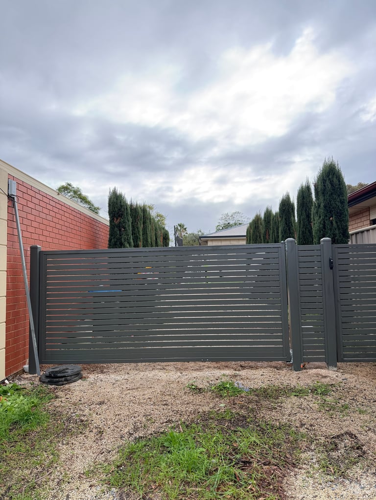 Dark gray horizontal slat gate with brick building and tall cypress trees in background under cloudy sky