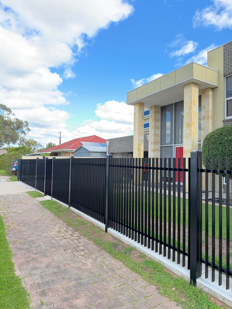Modern two-story house with cream and black exterior behind black metal fence on paved pathway, blue sky with clouds above
