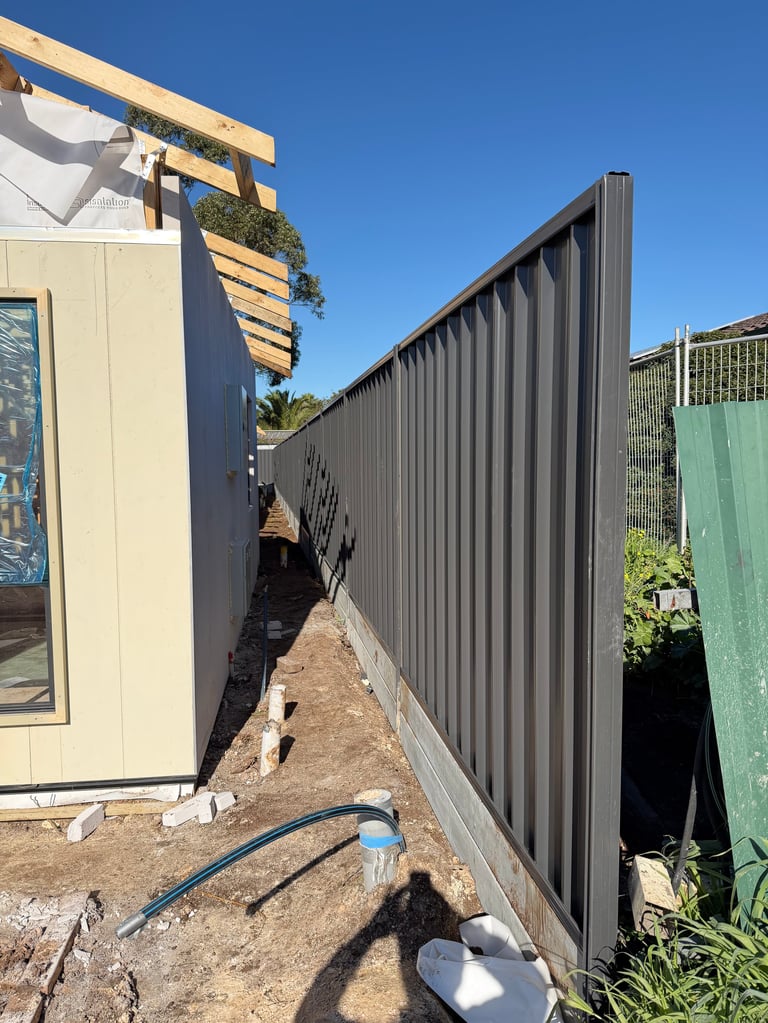 Narrow side yard between cream-colored residential structure and dark corrugated metal fence, with blue water hose on dirt ground
