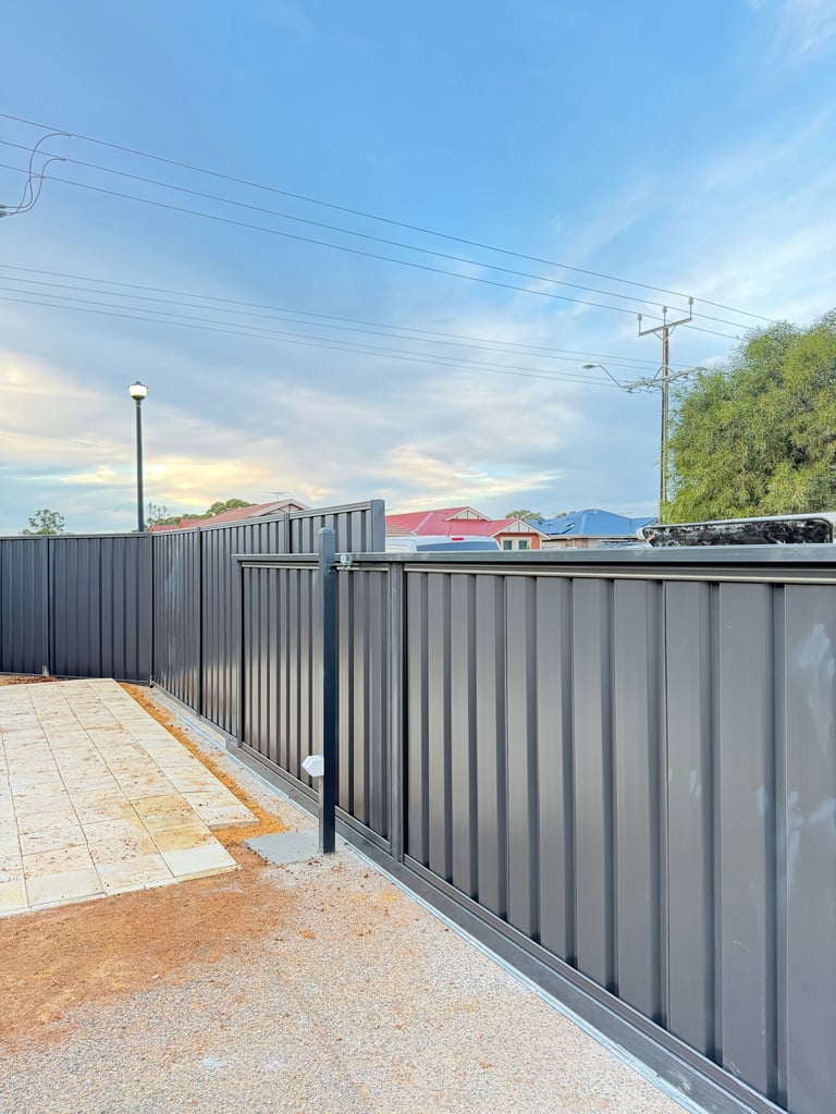 Metal fence with black vertical slats bordering a paved pathway against a blue sky with industrial buildings in background