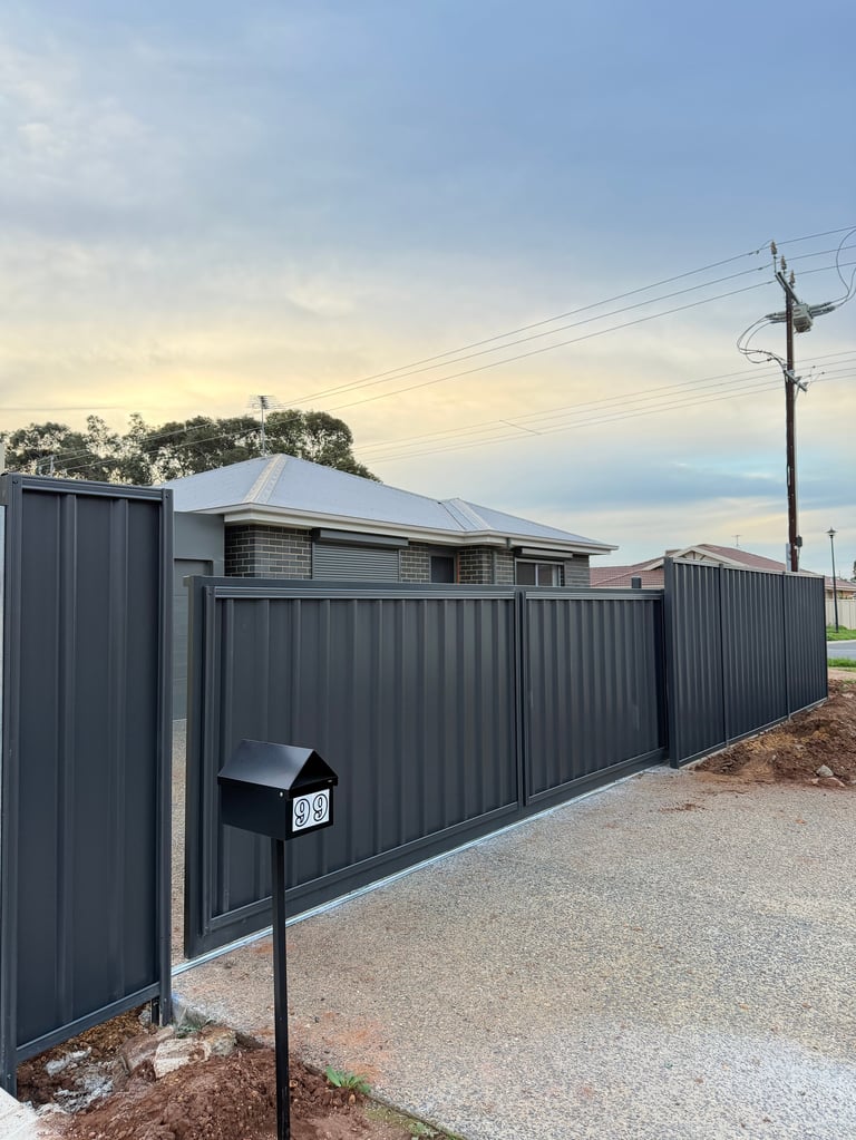 Modern house with corrugated metal fence and driveway, surrounded by trees and power lines at dusk