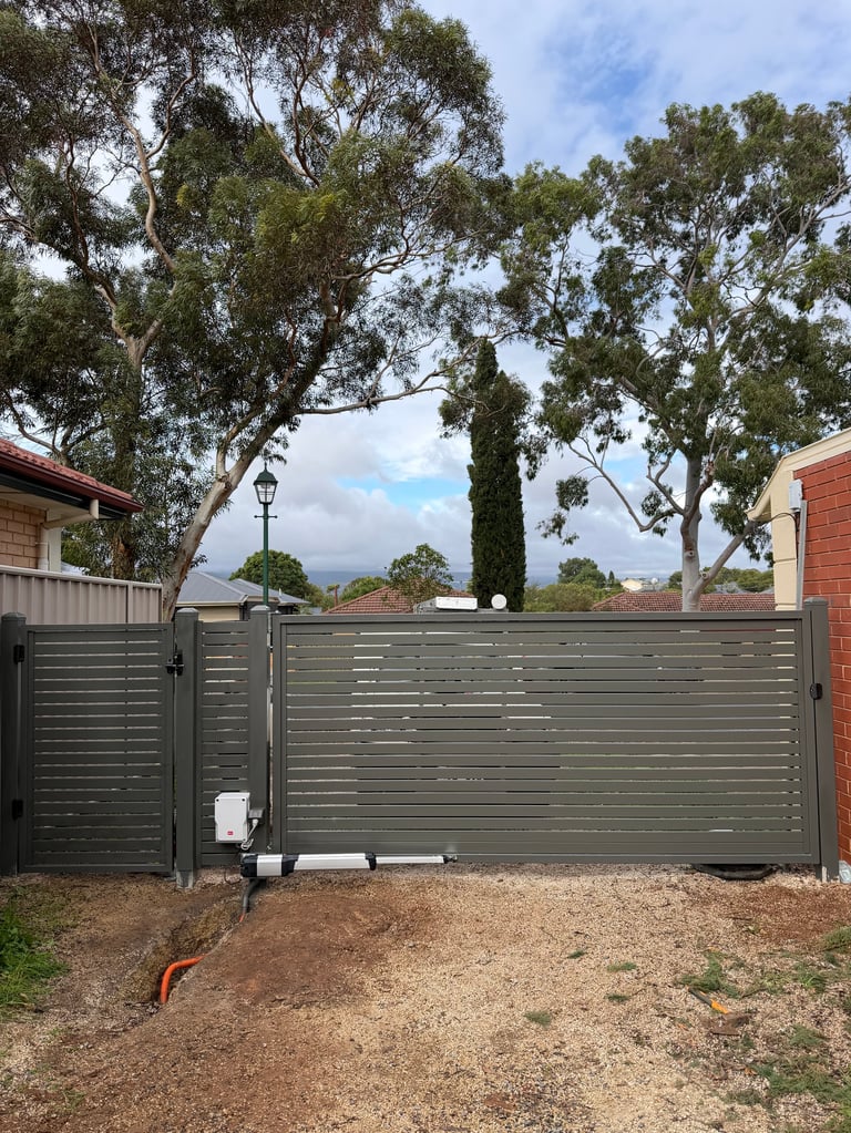 Modern dark green metal driveway gate with horizontal slats in a residential property with tall eucalyptus trees and mountains visible in background
