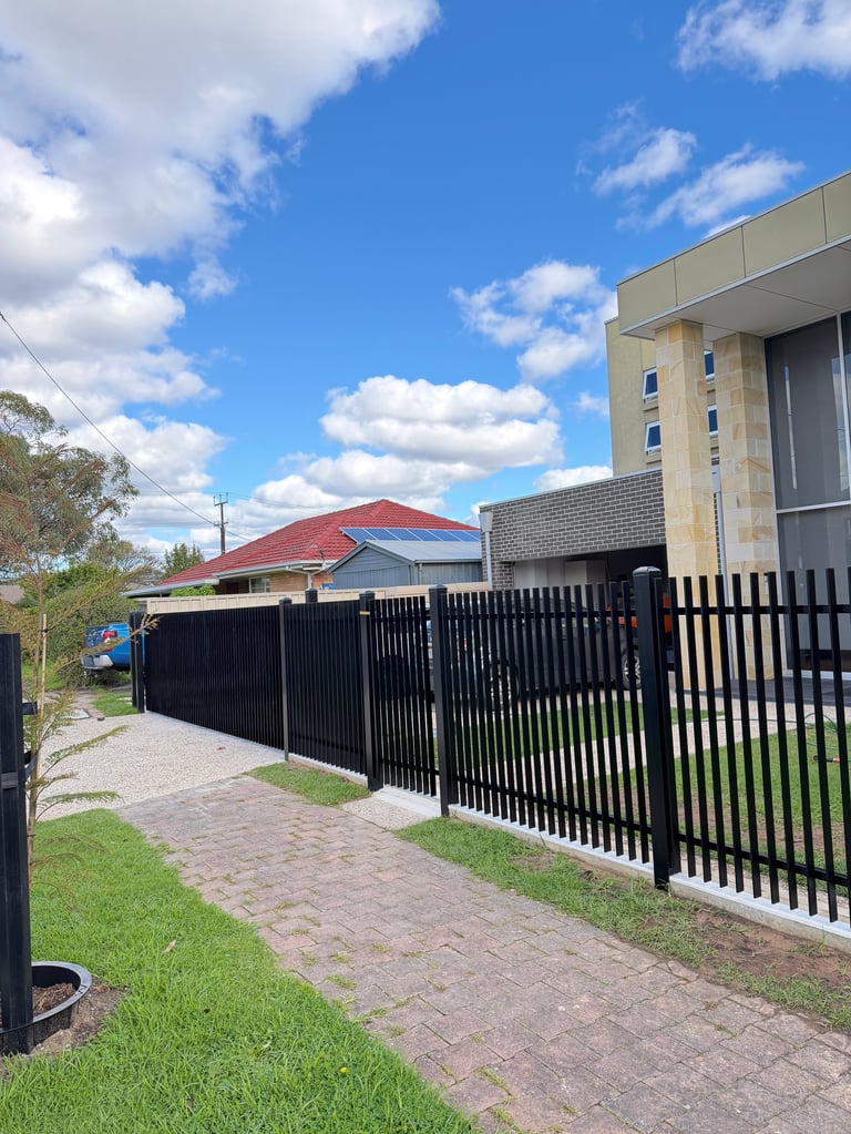 Modern residential property with black metal fence, paved driveway, and multi-story beige house under blue sky
