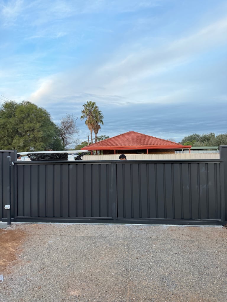 A modern dark gray metal sliding gate with a driveway, palm trees and a red-roofed building visible beyond it under a blue sky.