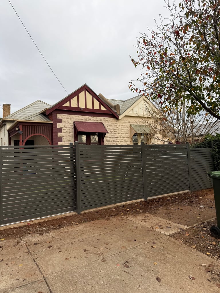 Modern dark green horizontal slat fence in front of a two-story house with red and cream trim and timber details
