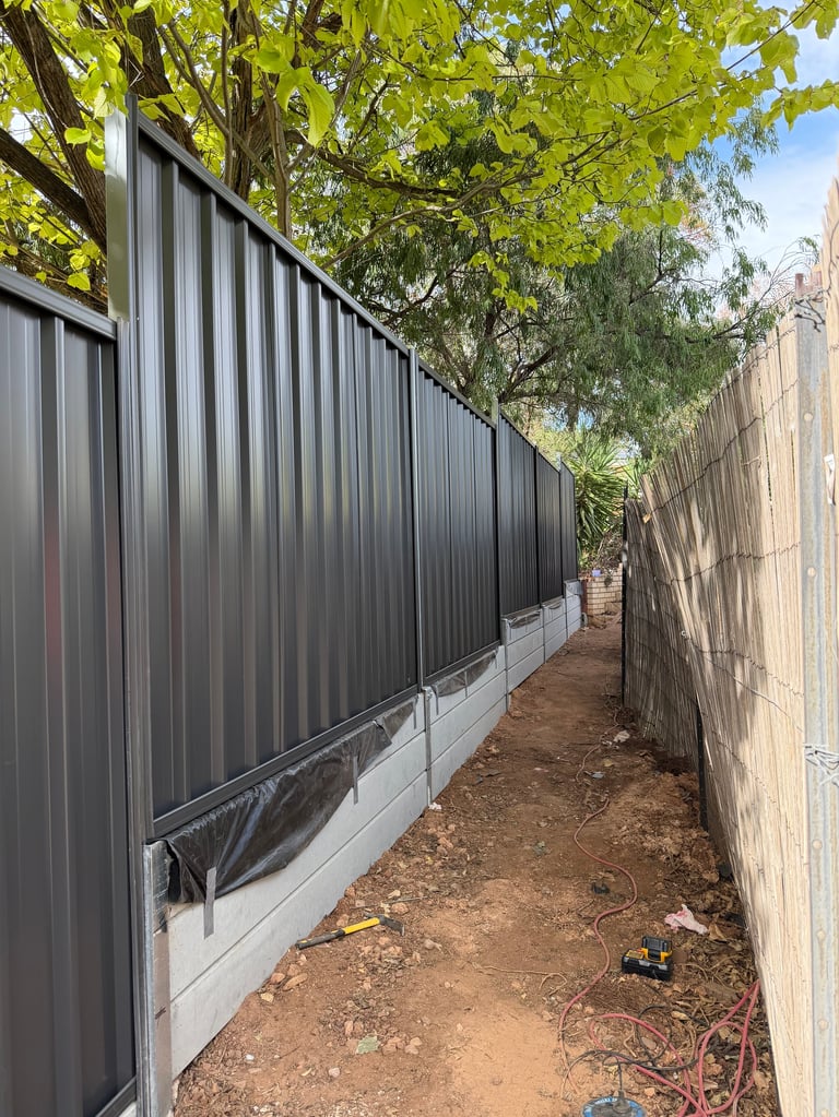 Narrow alley between corrugated metal fence and brick wall with dirt ground and large green trees overhead