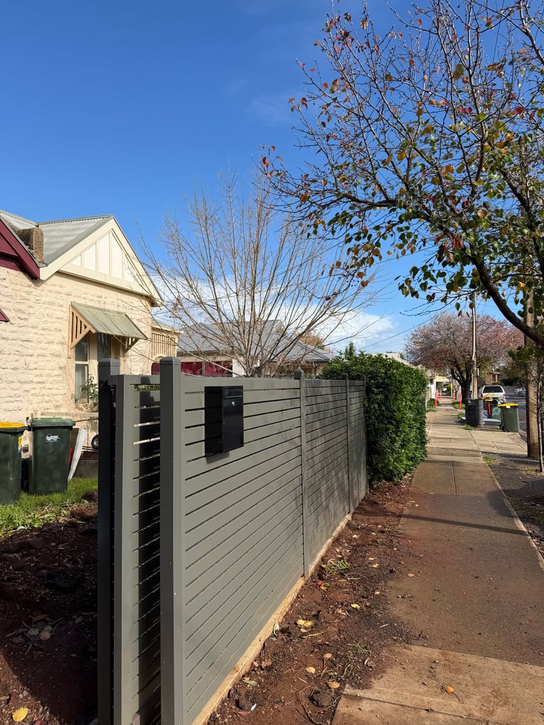 Residential street with grey horizontal slat fence, brick house, leafless tree, and clear blue sky