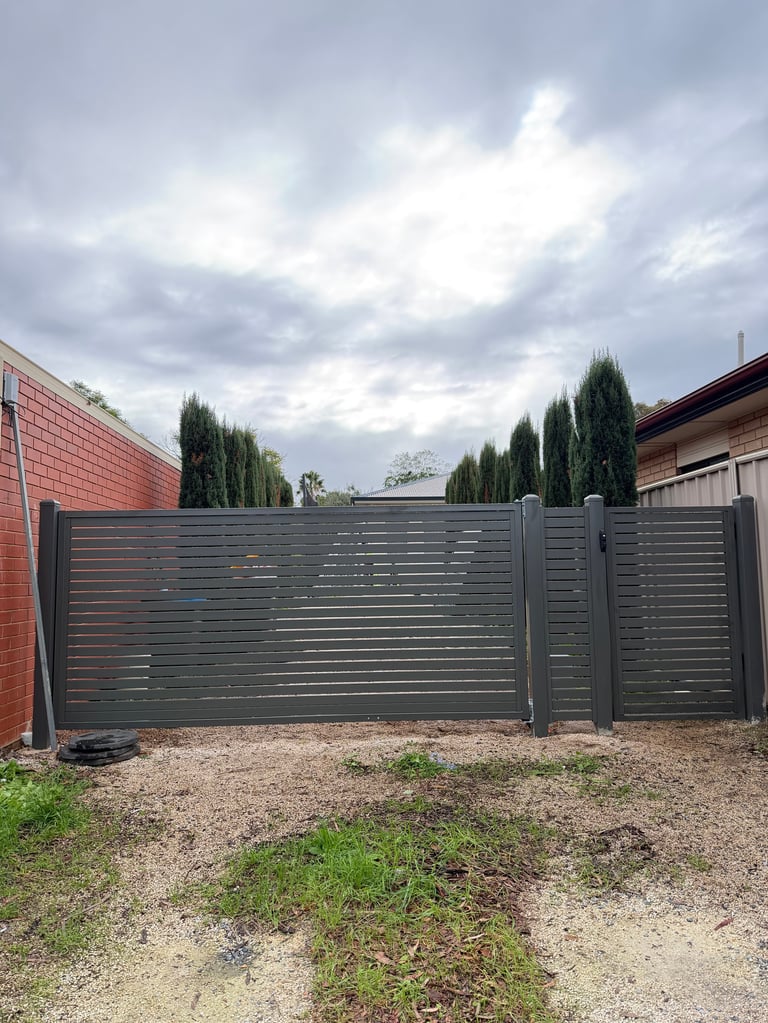 Modern gray horizontal slat driveway gate with tall cypress trees and brick wall under cloudy sky